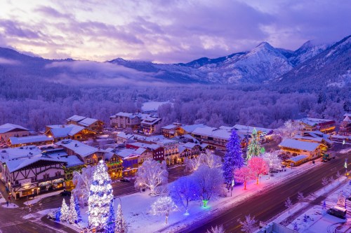 A snowy village illuminated with colorful lights, surrounded by mountains under a twilight sky.
