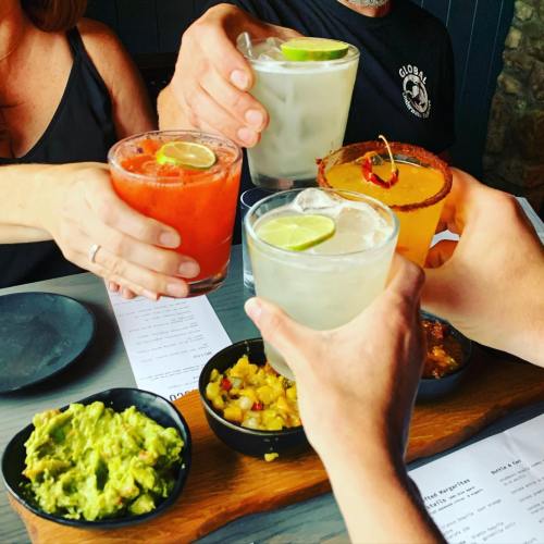 A group toasts with colorful cocktails, accompanied by bowls of guacamole and salsa on a restaurant table.