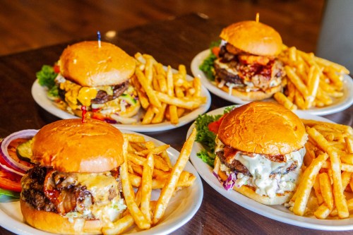 Four gourmet burgers with fries on white plates, featuring various toppings and sauces, served in a casual dining setting.