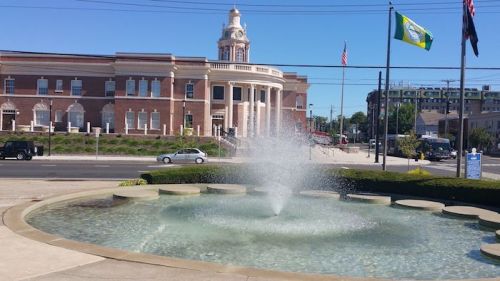 A fountain in the foreground with a historic building and flags in the background on a sunny day.
