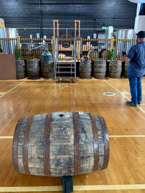 A wooden barrel in the foreground with a stage and barrels in the background, set in a spacious indoor venue.