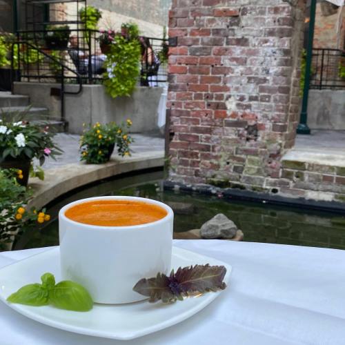 A white bowl of orange soup garnished with basil sits on a table near a brick wall and a tranquil water feature.