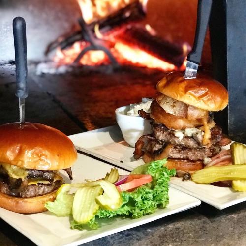 Two gourmet burgers on plates, one stacked high with toppings, accompanied by pickles, lettuce, and a side of sauce.