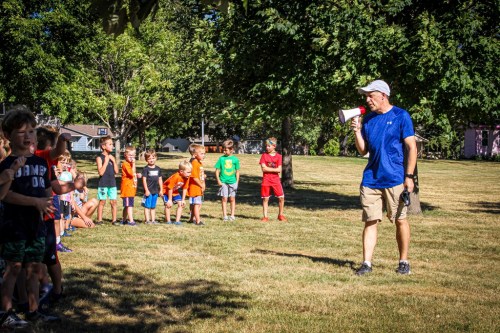 A coach addresses a line of children in a park, holding a megaphone, with trees and grass in the background.