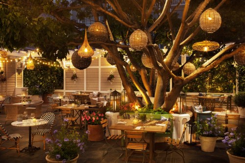 A cozy outdoor restaurant with string lights, woven lanterns, and tables surrounded by greenery and flowers.
