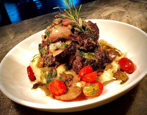 A plate of tender beef topped with herbs, served on creamy mashed potatoes with cherry tomatoes and a rich sauce.