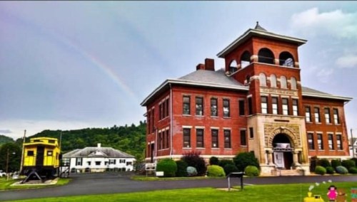 Historic red brick building with a clock tower, surrounded by greenery and a rainbow in the sky.