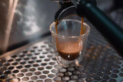A close-up of espresso being brewed into a clear glass cup on a metal surface with circular holes.