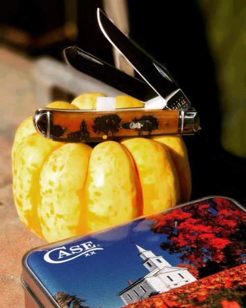 A pocket knife rests on a yellow pumpkin, next to a tin featuring a church and autumn scenery.