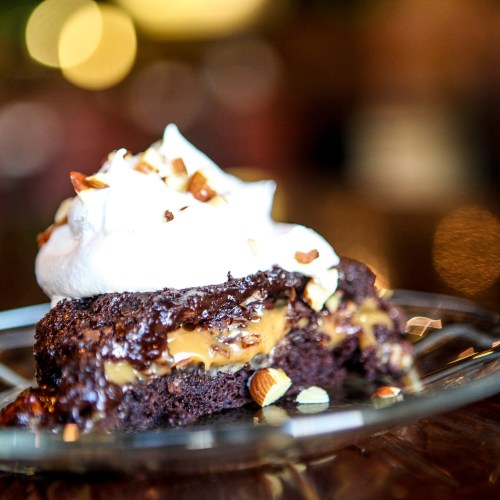A close-up of a chocolate dessert with caramel filling, topped with whipped cream and chopped nuts on a glass plate.