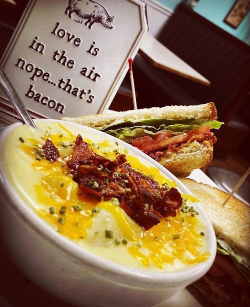 A bowl of cheesy potato soup topped with bacon, alongside a sandwich and a humorous sign about love and bacon.