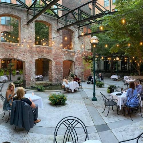 Outdoor dining area with string lights, tables set for guests, and greenery in a charming brick courtyard.
