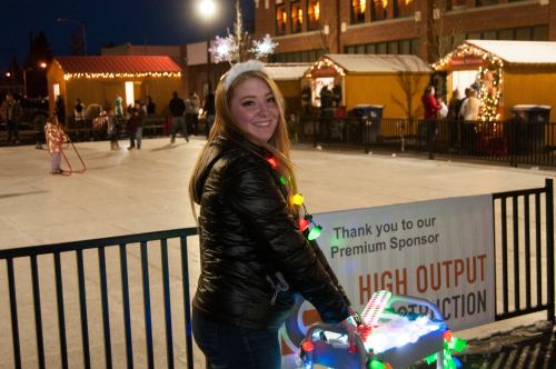 A smiling woman in a festive outfit with lights and a snowflake headband stands near an ice skating rink at night.