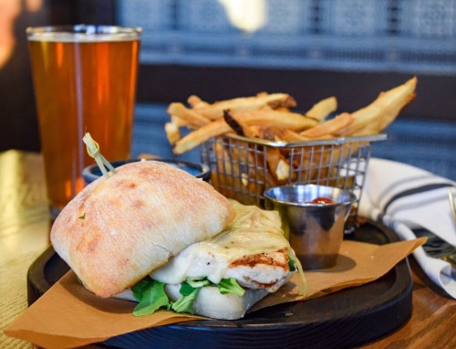 A grilled chicken sandwich with cheese on a plate, served with fries and a pint of beer in the background.