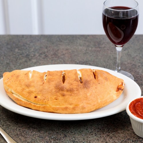 A golden-brown calzone on a white plate, accompanied by marinara sauce and a glass of red wine.