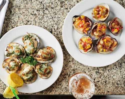 A plate of baked oysters with toppings, alongside a plate of stuffed clams, with a lemon wedge and a glass of beer.