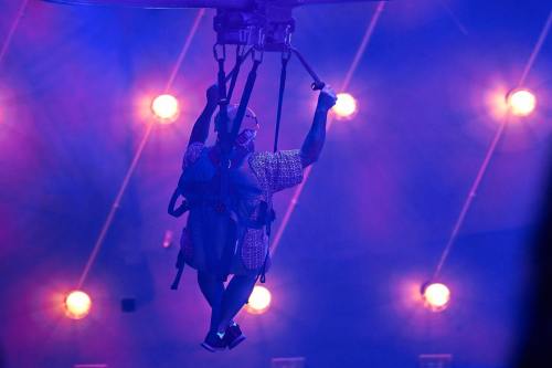 A person in a harness is zip-lining through a colorful, illuminated space with bright lights in the background.