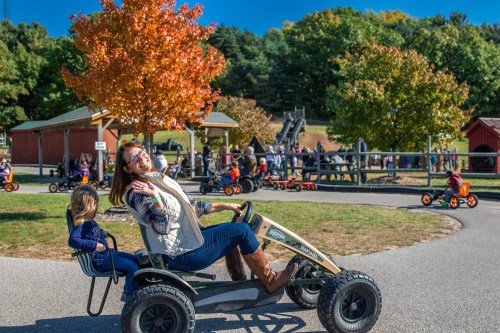 A woman waves while riding a pedal cart with a child in a park filled with autumn trees and families enjoying the day.