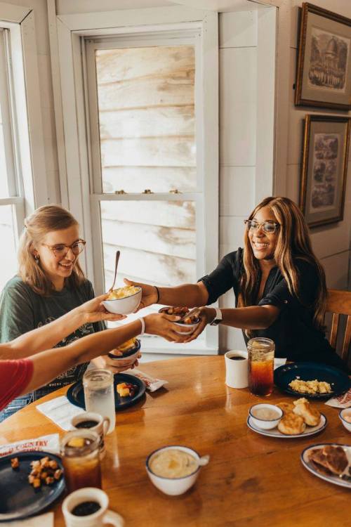 Two women share food at a table, smiling and enjoying a meal together, with drinks and plates of food around them.