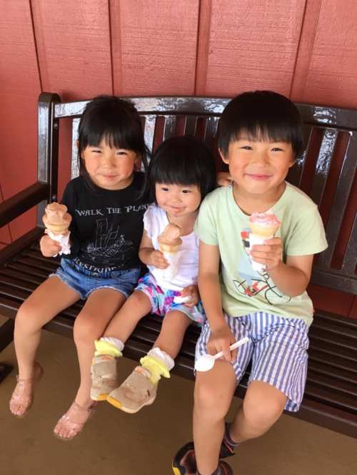 Three children sitting on a bench, happily holding ice cream cones, smiling at the camera.