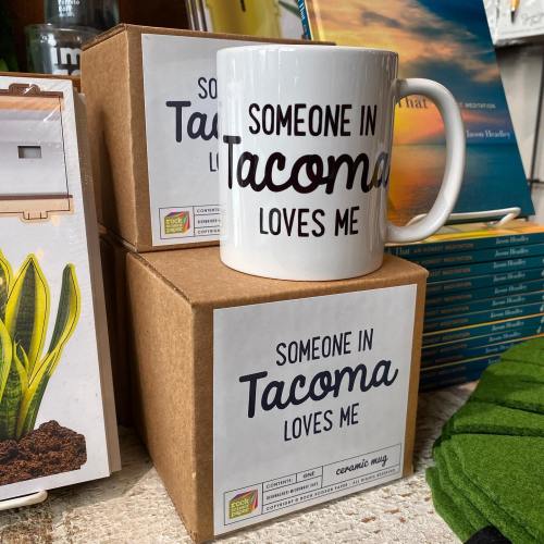 A white mug with "Someone in Tacoma loves me" printed on it, placed next to two brown boxes in a cozy setting.