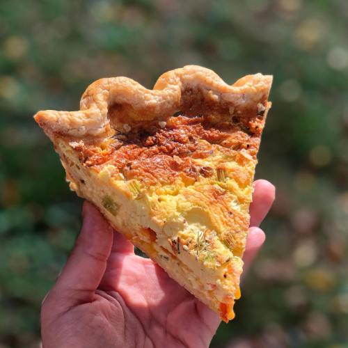 A hand holding a slice of quiche with a golden crust and colorful filling, set against a blurred outdoor background.