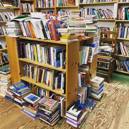 A wooden bookshelf overflowing with books, some stacked on the floor, in a cozy, cluttered bookstore.
