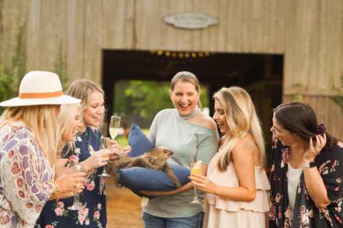 A group of smiling women gather around a woman holding a sloth, celebrating with drinks in a rustic outdoor setting.