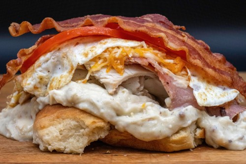 A hearty sandwich with layers of bacon, eggs, cheese, ham, and creamy sauce on a biscuit, garnished with tomato slices.