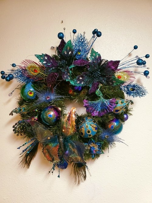 A vibrant wreath adorned with peacock feathers, colorful ornaments, and lush greenery, set against a light wall.
