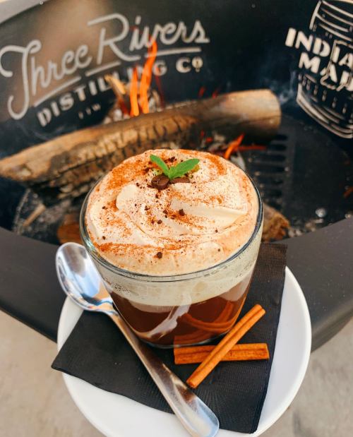 A glass of coffee topped with whipped cream and a sprinkle of cocoa, garnished with a mint leaf and cinnamon sticks.