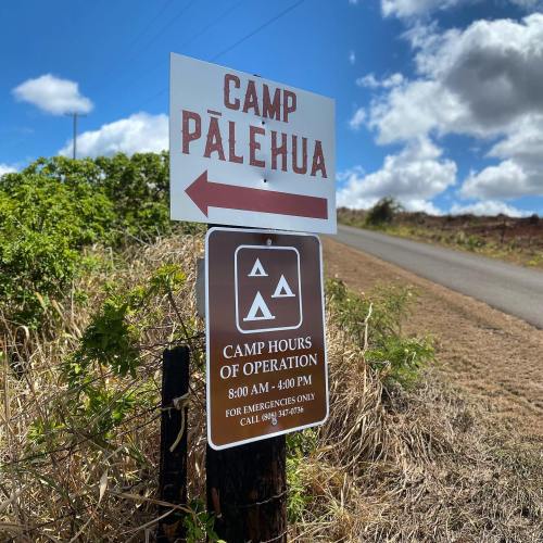 Sign for Camp Pālehua with hours of operation and emergency contact information, located along a rural road.