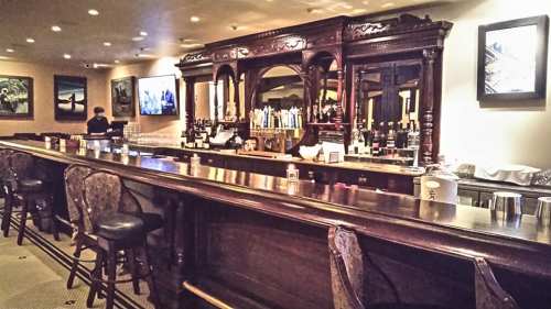 A wooden bar with high stools, bottles on shelves, and artwork on the walls in a cozy, well-lit setting.