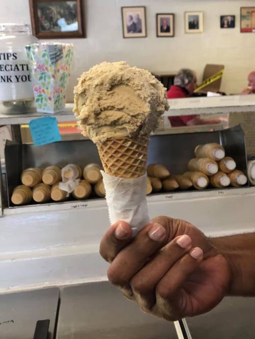 A hand holding a large scoop of brown ice cream on a cone, with a background of ice cream containers and people.