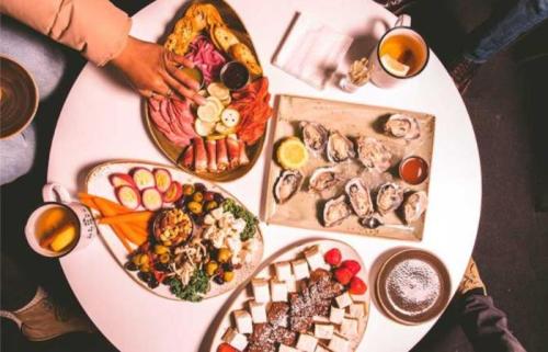 A table spread with various dishes, including charcuterie, oysters, salads, and desserts, surrounded by drinks.