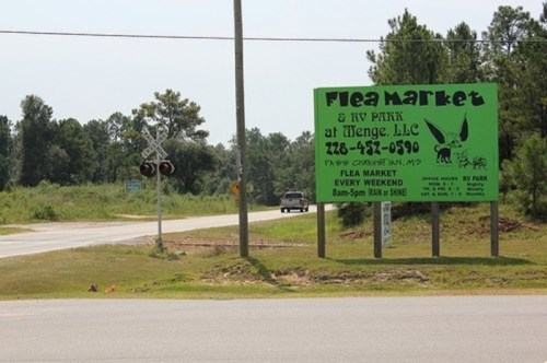A green sign for a flea market and RV park, with contact info, near a road and railroad crossing in a rural area.