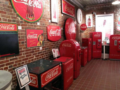 A vintage Coca-Cola display featuring red vending machines, signs, and memorabilia against a brick wall.