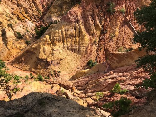 A colorful canyon with layers of red, yellow, and orange rock, surrounded by sparse greenery.