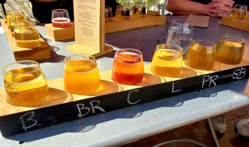 A flight of craft beers in various colors served in small glasses on a wooden tray, labeled with initials.