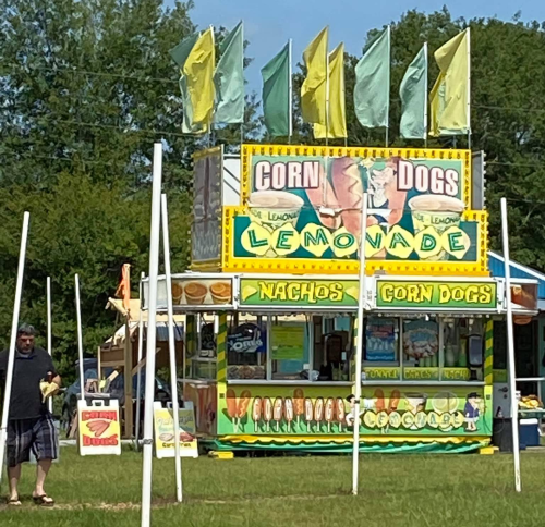 A colorful food stand at a fair selling lemonade, corn dogs, and nachos, with flags waving in the background.