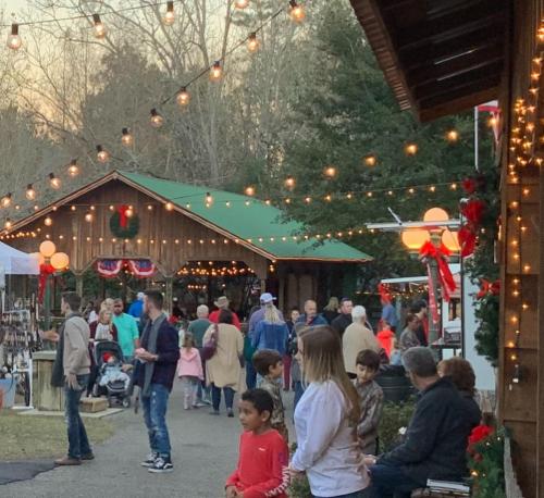 A festive outdoor market scene with people shopping, holiday decorations, and string lights in the evening.