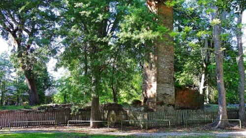 Ruins of a brick structure surrounded by tall trees and greenery, with a wooden fence in the foreground.