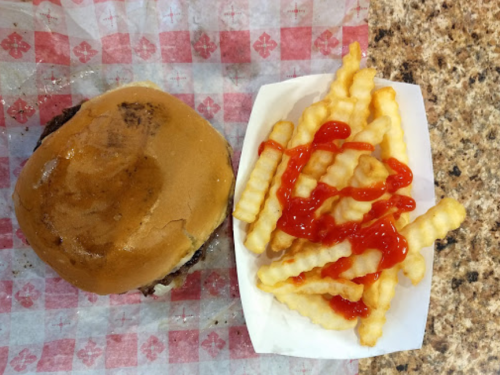 A hamburger on a red and white checkered paper, alongside a serving of crinkle-cut fries drizzled with ketchup.