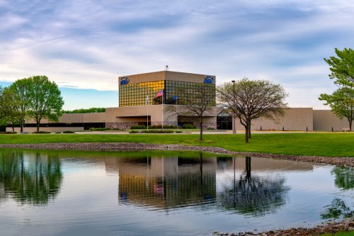 Modern building with large glass windows, surrounded by greenery and a pond reflecting the structure.