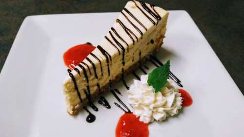 A slice of cheesecake drizzled with chocolate, served with strawberry sauce and whipped cream on a white plate.