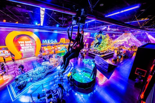 A vibrant indoor amusement park with colorful lights, games, and attractions, featuring a person zip-lining above.