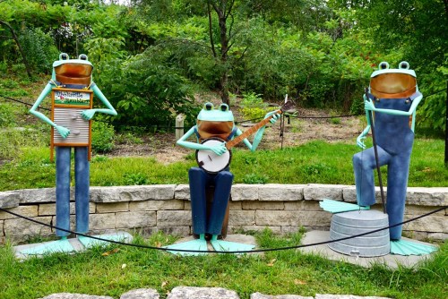 Three whimsical frog sculptures playing musical instruments in a green outdoor setting.