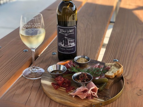 A wooden platter with assorted meats, pickles, and bread, alongside a glass of white wine and a bottle on a table.