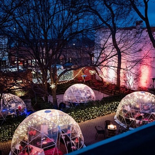 A cozy outdoor dining area with illuminated igloo-like structures surrounded by trees and festive lights at dusk.