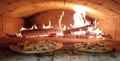 Two pizzas baking in a wood-fired oven, with flames and logs visible in the background.